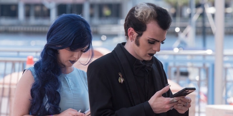 Un hombre y una mujer con peinados llamativos y maquillaje oscuro miran sus teléfonos.