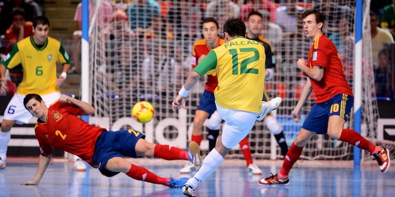Un jugador de fútbol sala patea la pelota hacia el arco contrario mientras otros jugadores intentan interceptarlo.
