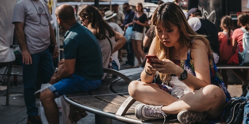 Una mujer observa su teléfono sentada en un banco en la ciudad, rodeada de gente.